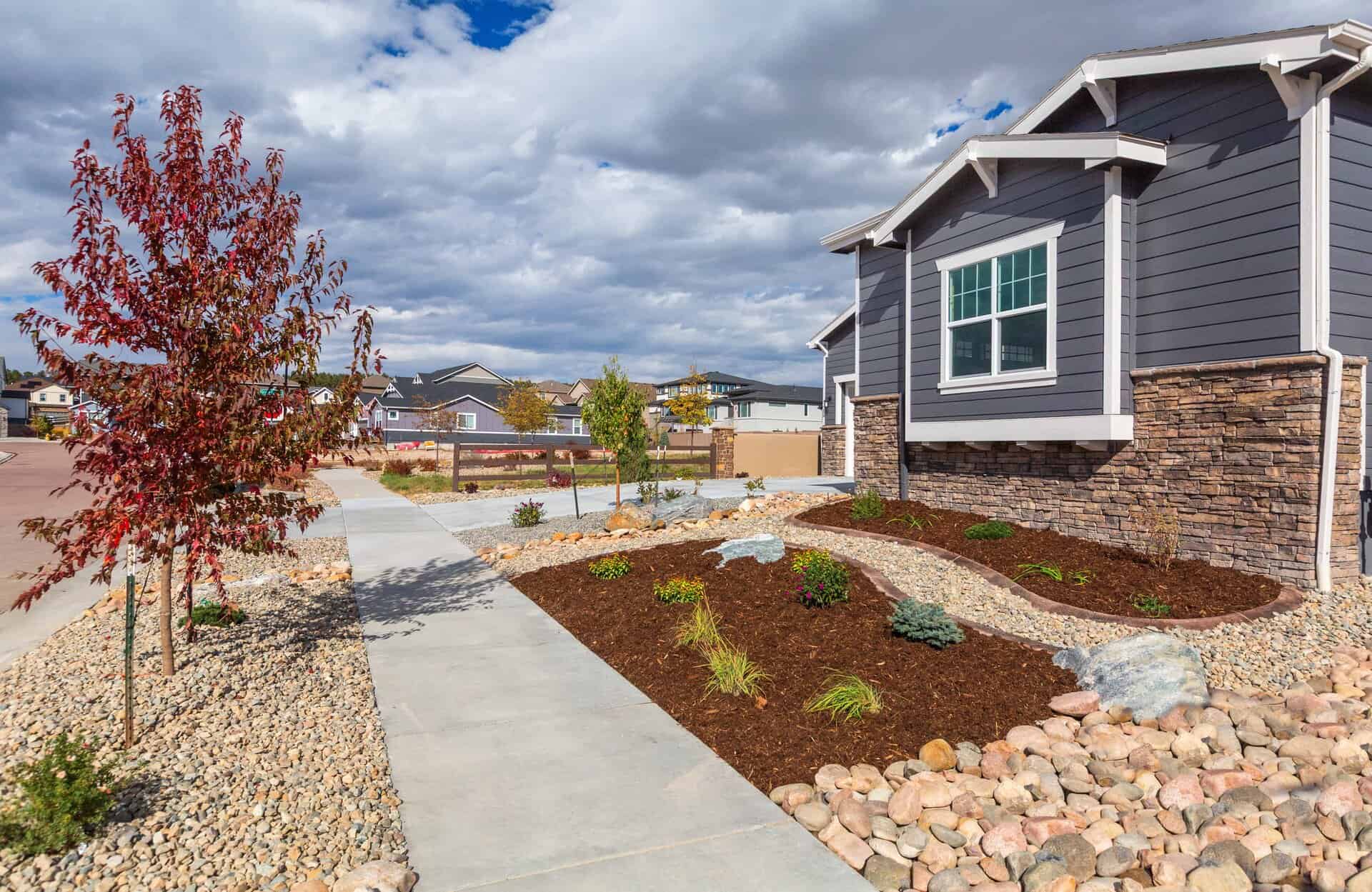 Front yard of a modern house designed by a skilled landscaper, featuring stone landscaping, small trees, shrubs, and a concrete walkway under a partly cloudy sky.