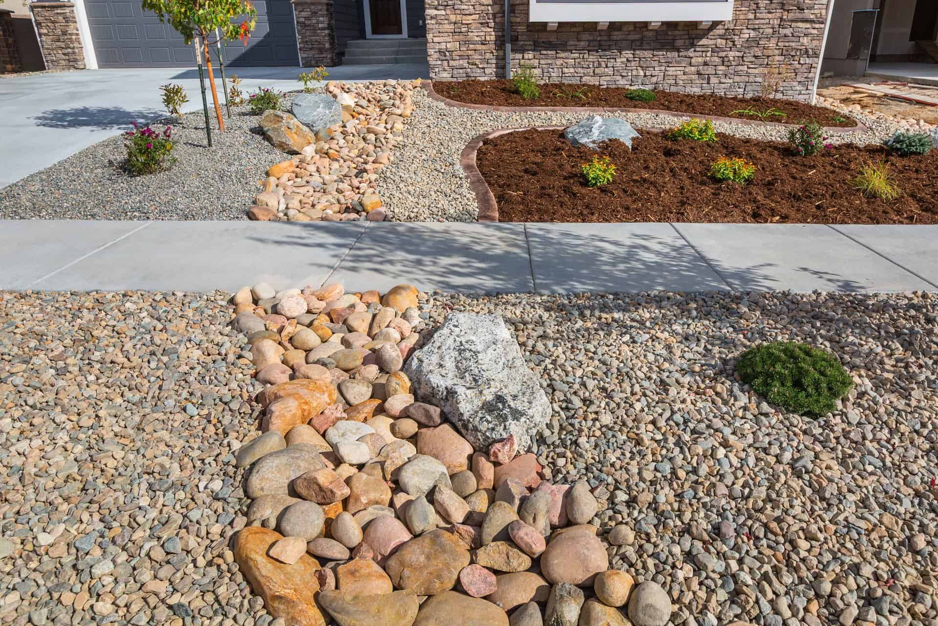 A skilled landscaper designed this front yard with a dry riverbed of large rocks, mulch planting beds with small shrubs, and a stone pathway leading to a charming brick house.