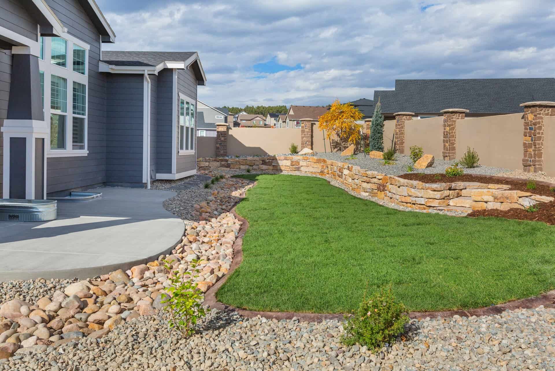 A modern house with a landscaped backyard, crafted by a skilled landscaper, features a green lawn, curved stone retaining walls, decorative rocks, and young plants under a partly cloudy sky.