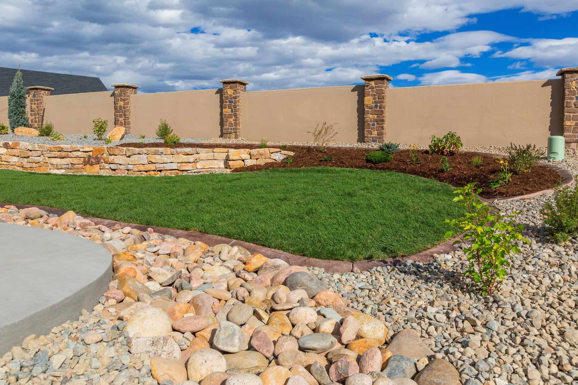 A landscaper-designed backyard features a green grass lawn, stone retaining wall, rock garden, and a beige privacy fence with brick pillars under a partly cloudy sky.