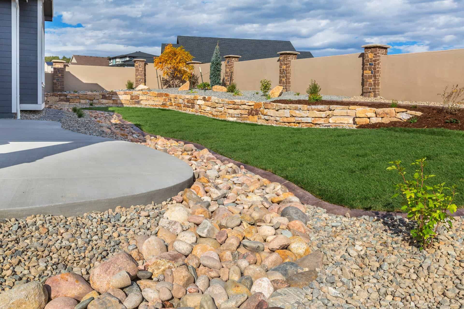 A landscaper-designed backyard features a curved concrete patio, a dry rock stream bed, green lawn, tiered stone retaining wall, young plants, and a beige fence with elegant stone pillars.