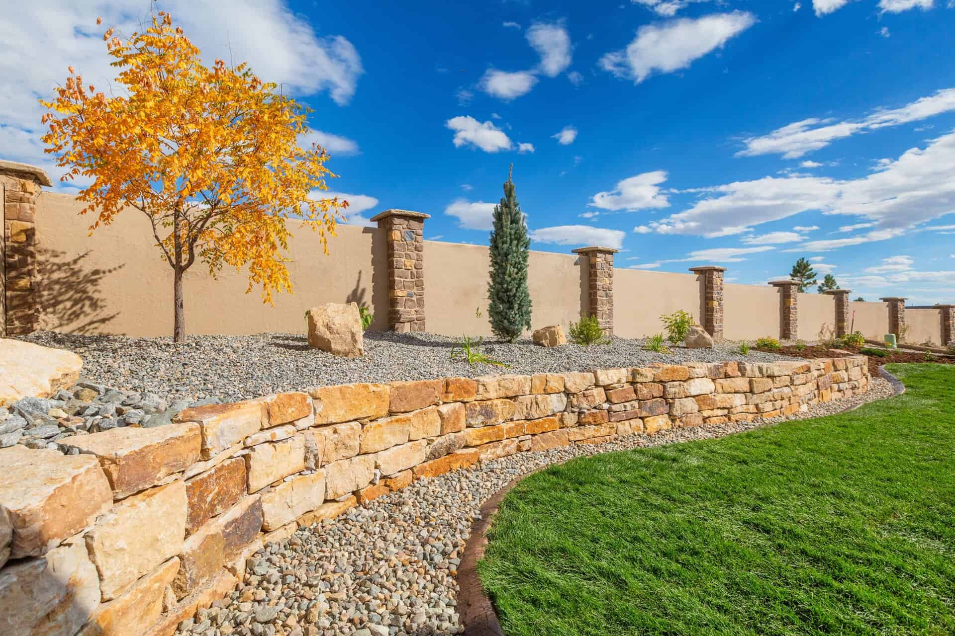 A landscaped backyard by a skilled landscaper features a stone retaining wall, gravel ground cover, a small tree with yellow leaves, and a green lawn under a blue sky with scattered clouds.