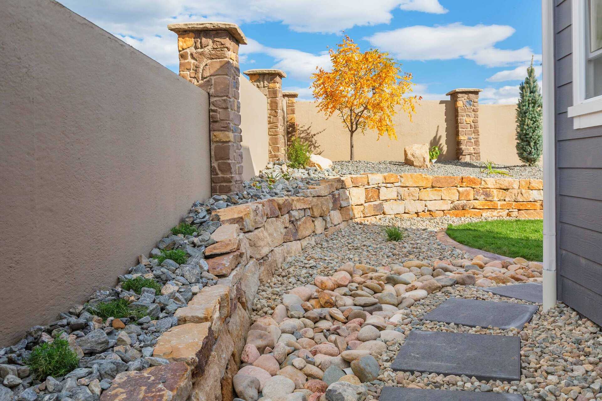 Stone path and rock landscaping by an expert landscaper curve along a retaining wall with a small tree, set against a beige privacy fence and blue sky with clouds.