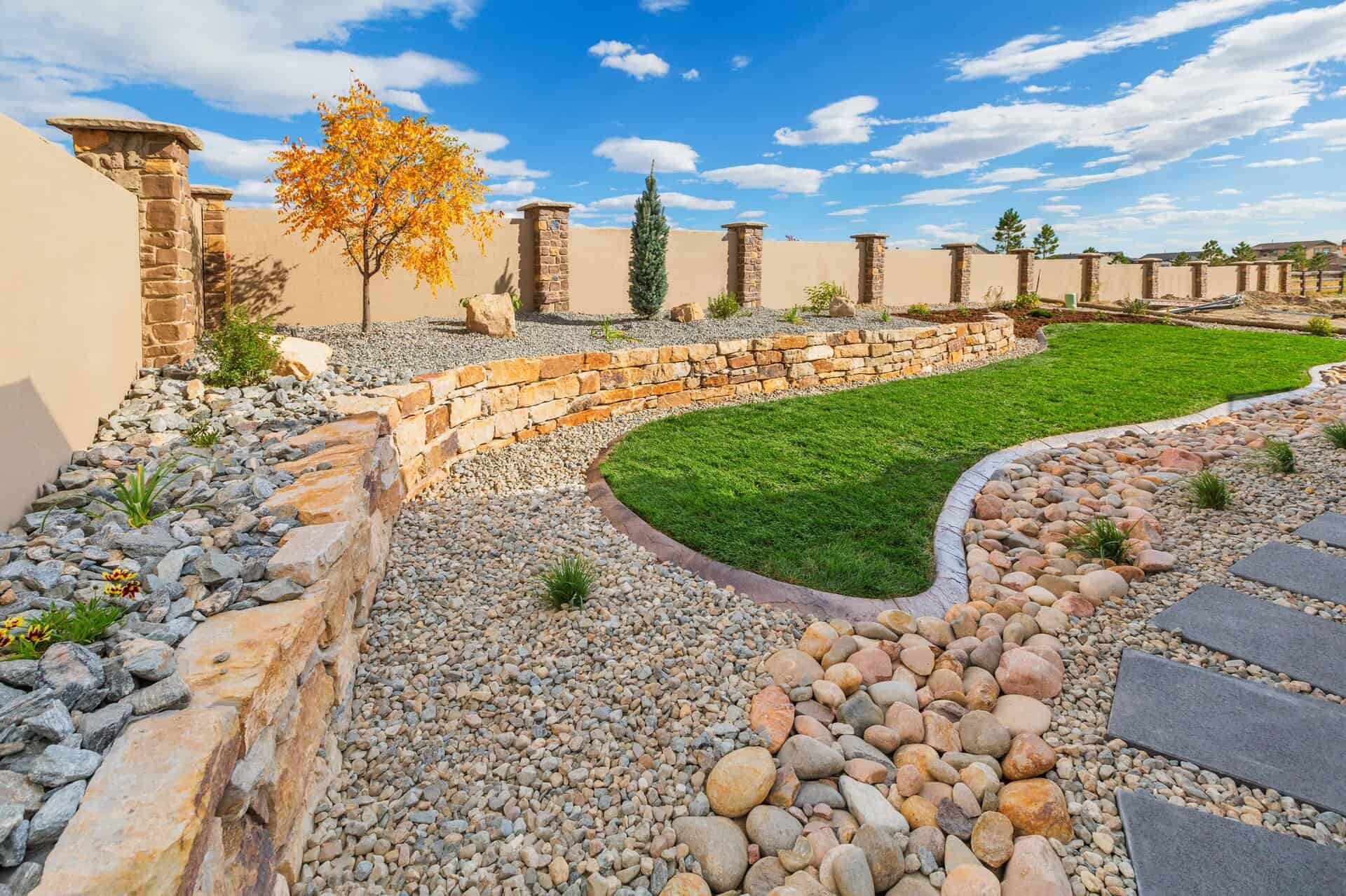 Landscaping Waterton Canyon Nielson Res_16 A landscaper-designed backyard with stone retaining walls, gravel beds, a small tree, lush green grass, and a stone pathway beneath a blue sky dotted with scattered clouds.