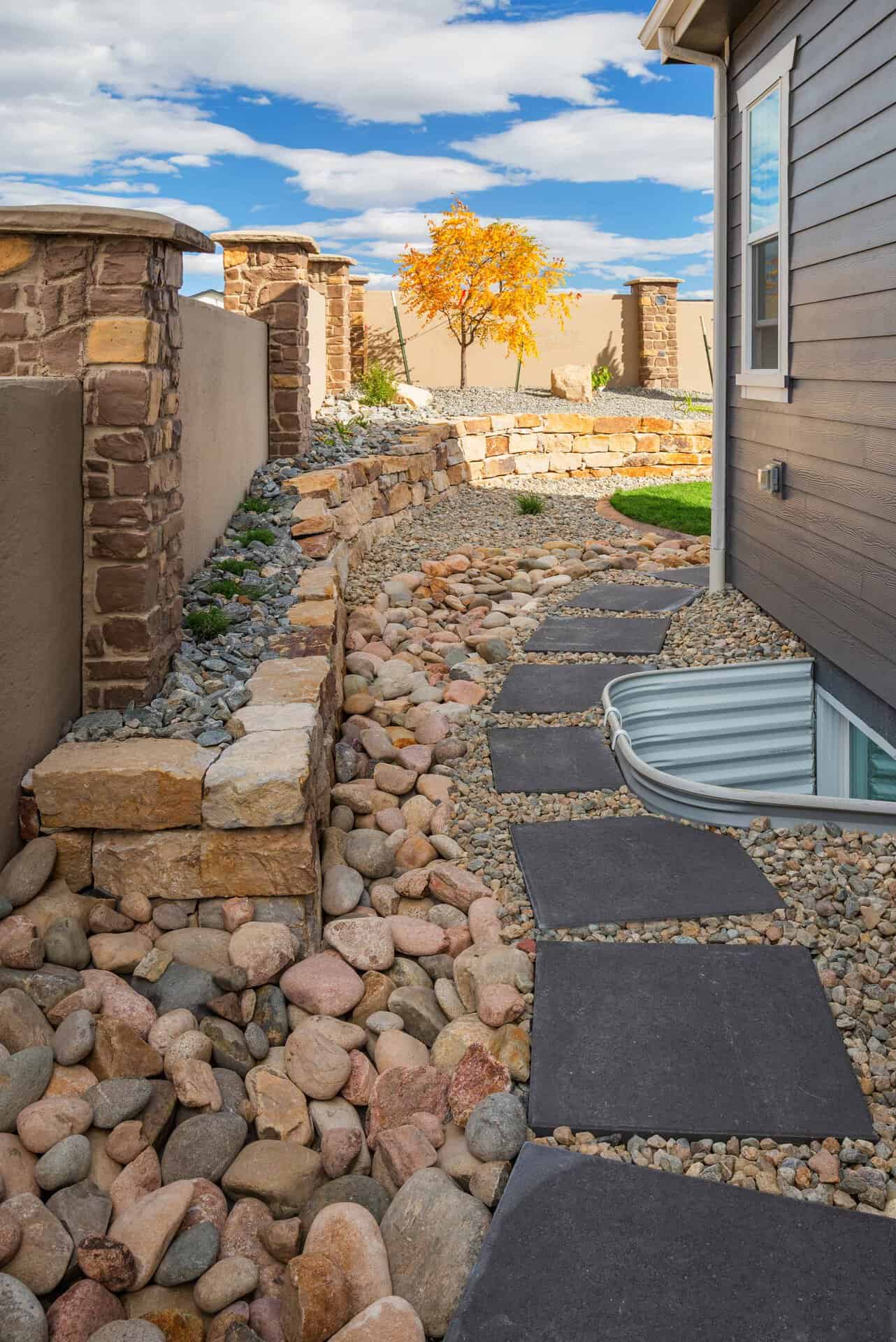 A stone pathway with large rocks, crafted by a skilled landscaper, leads alongside a house with dark siding, bordered by stone walls and a small tree with yellow leaves in the background.