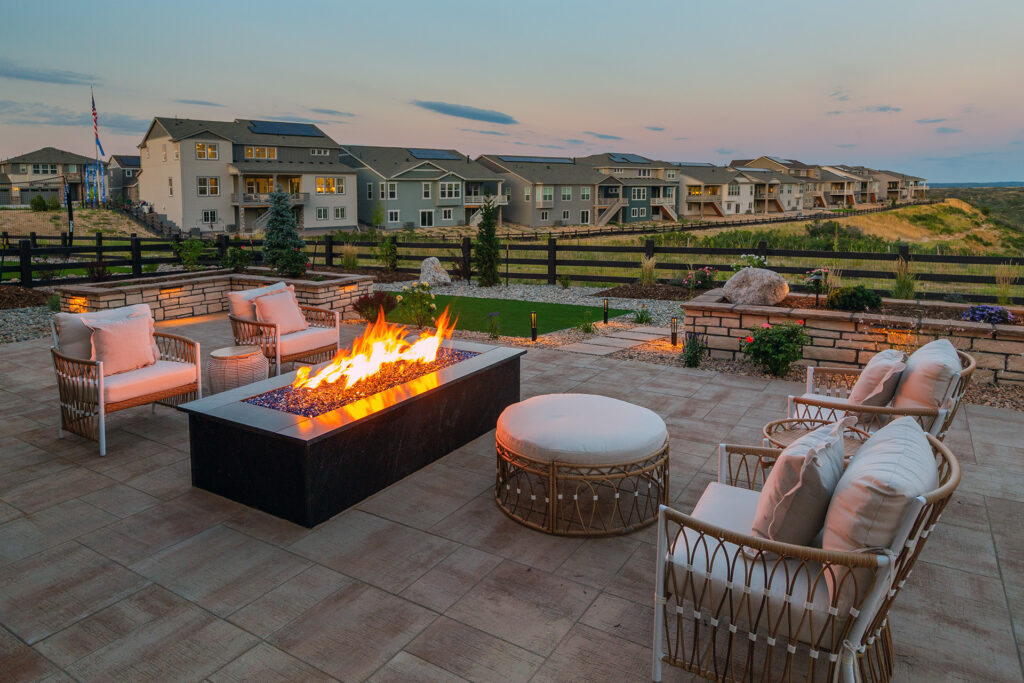 A modern outdoor patio with cushioned wicker chairs and a rectangular fire pit, overlooking houses and an open landscape at sunset.
