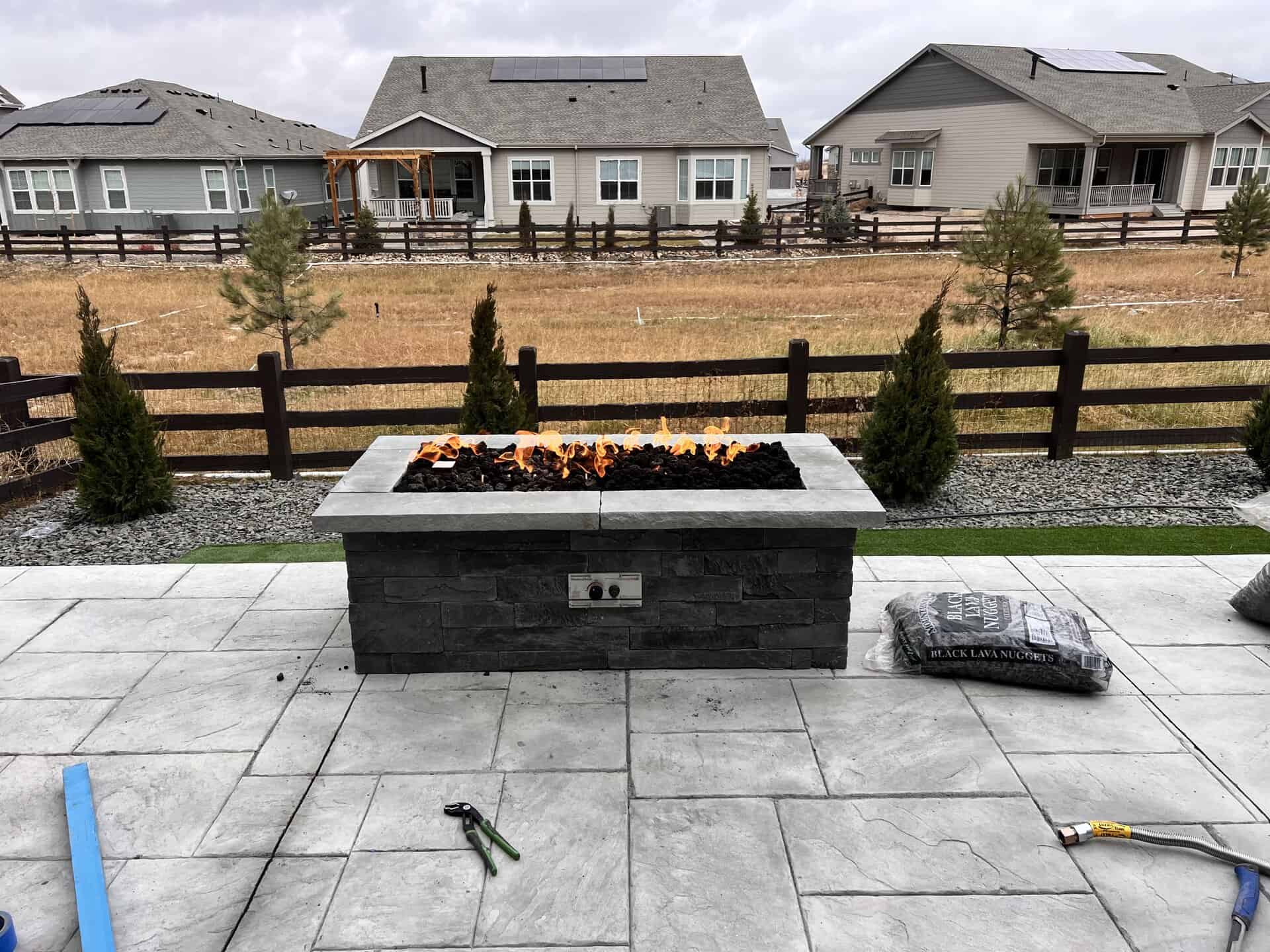A rectangular outdoor gas fire pit with flames is on a stone patio, surrounded by tools and bags, with houses and a fenced yard in the background.