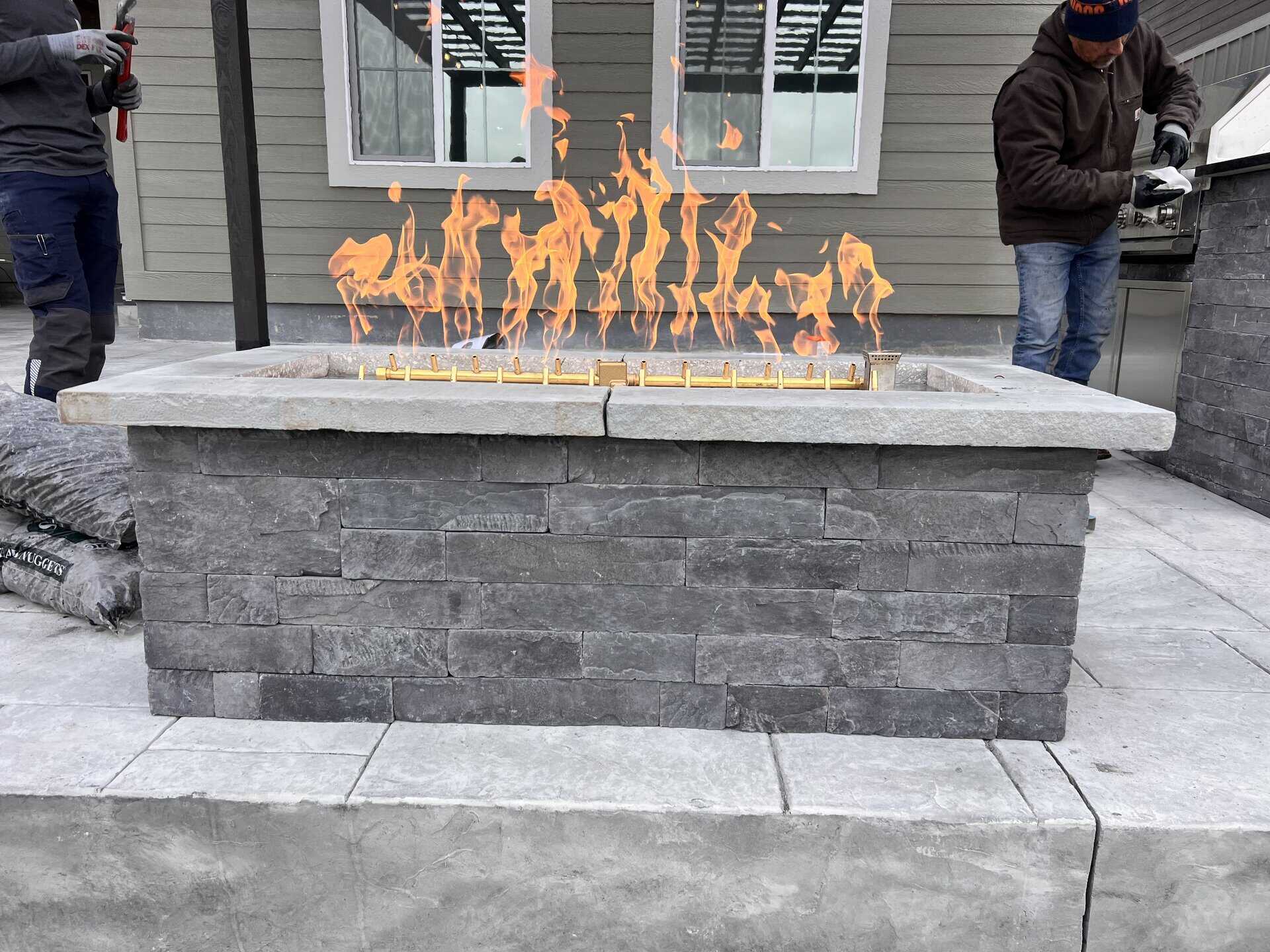 A rectangular stone fire pit with visible flames, set on a paved patio, with two people working nearby.