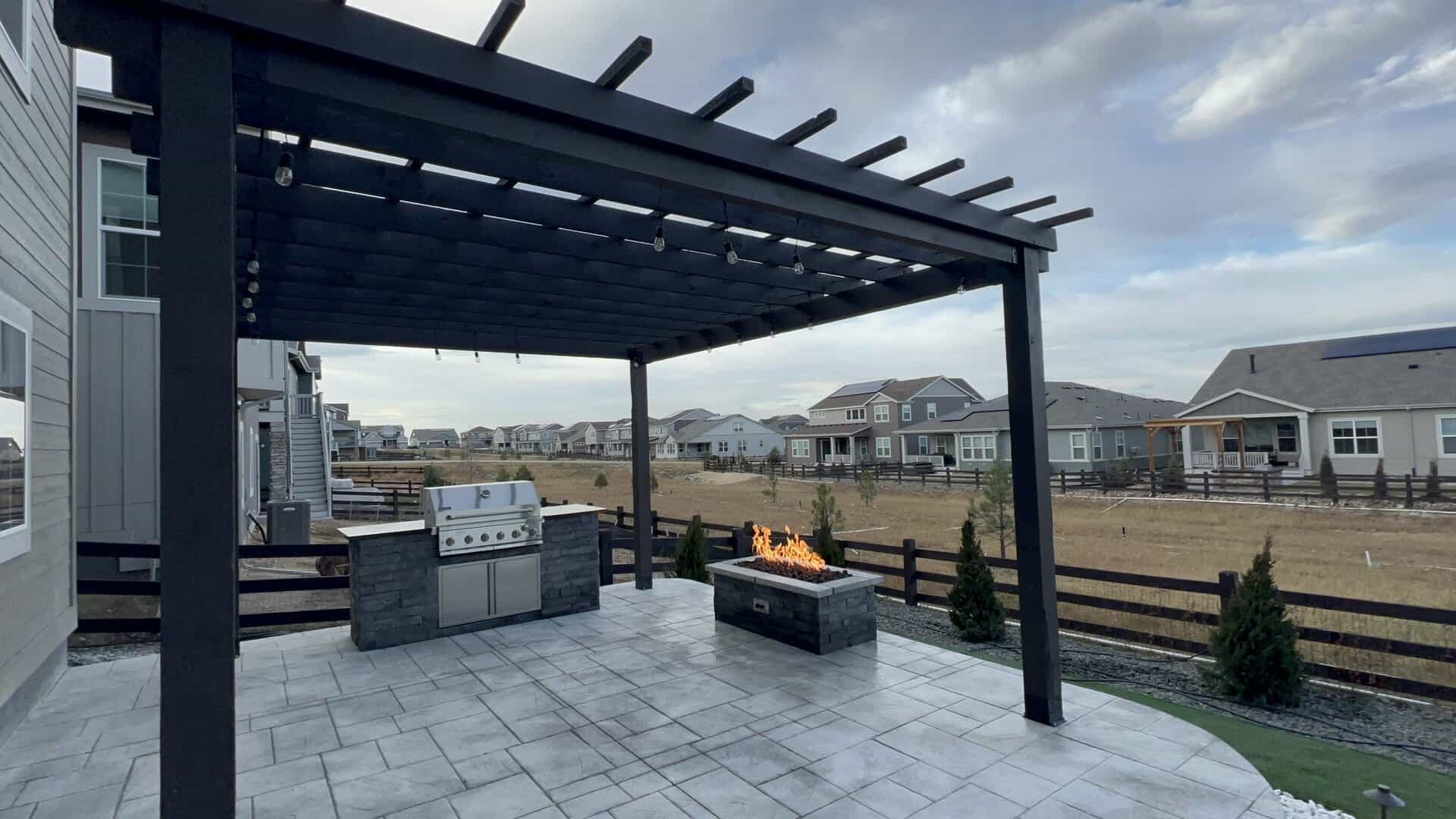 Modern backyard patio with a black pergola, built-in grill, and rectangular fire pit, overlooking a suburban neighborhood with houses and a fenced yard.