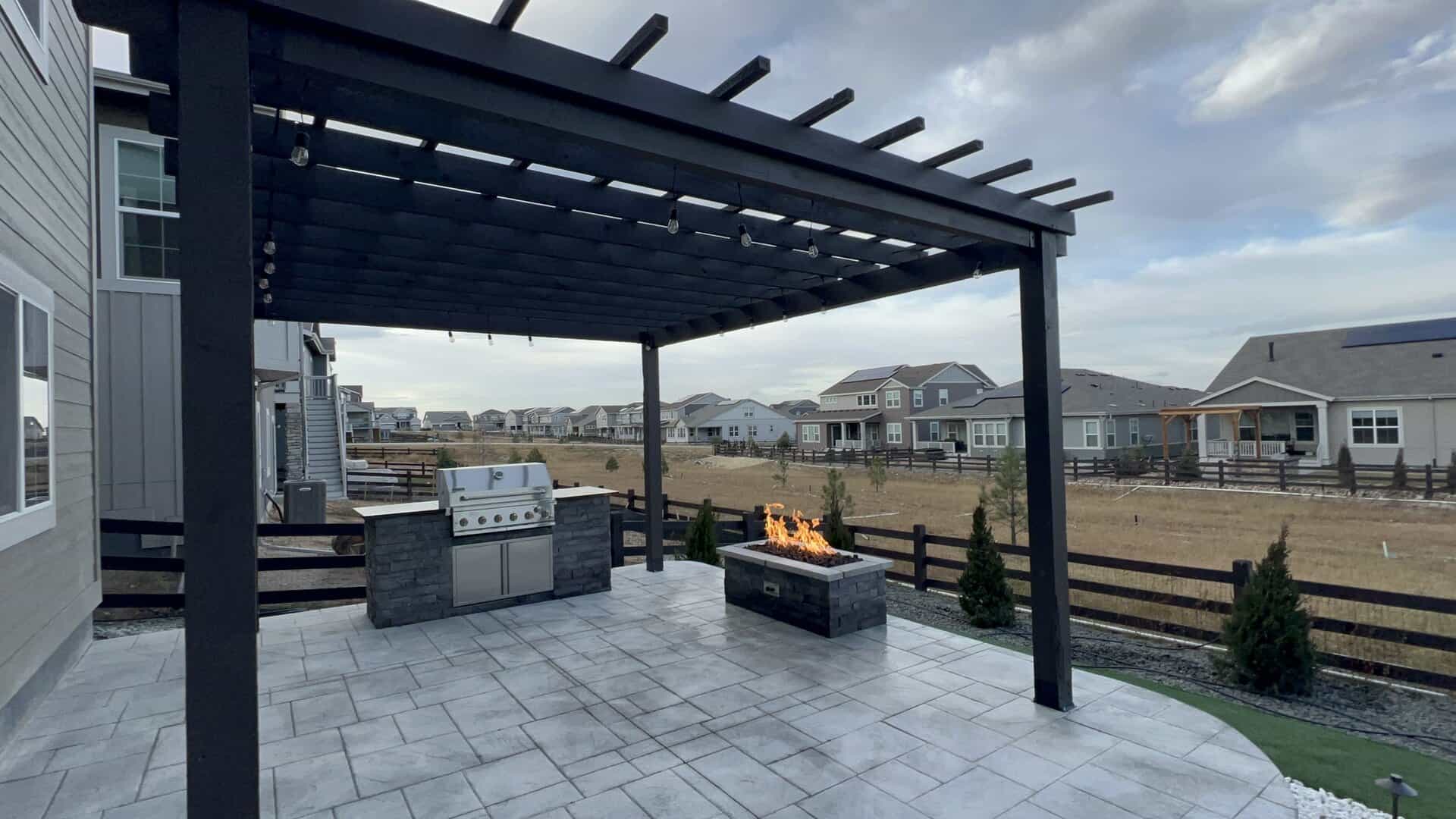 A modern patio with a pergola, built-in grill, and rectangular fire pit overlooks a fenced suburban neighborhood with houses and open land.