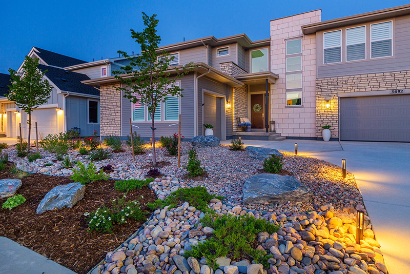 Modern two-story house with stone accents, landscaped front yard with rocks and small plants, illuminated by outdoor lighting at dusk.