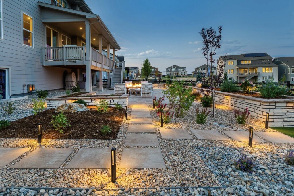 Modern backyard with a stone pathway, garden beds, outdoor dining area, and illuminated pathway lights in a suburban neighborhood at dusk.