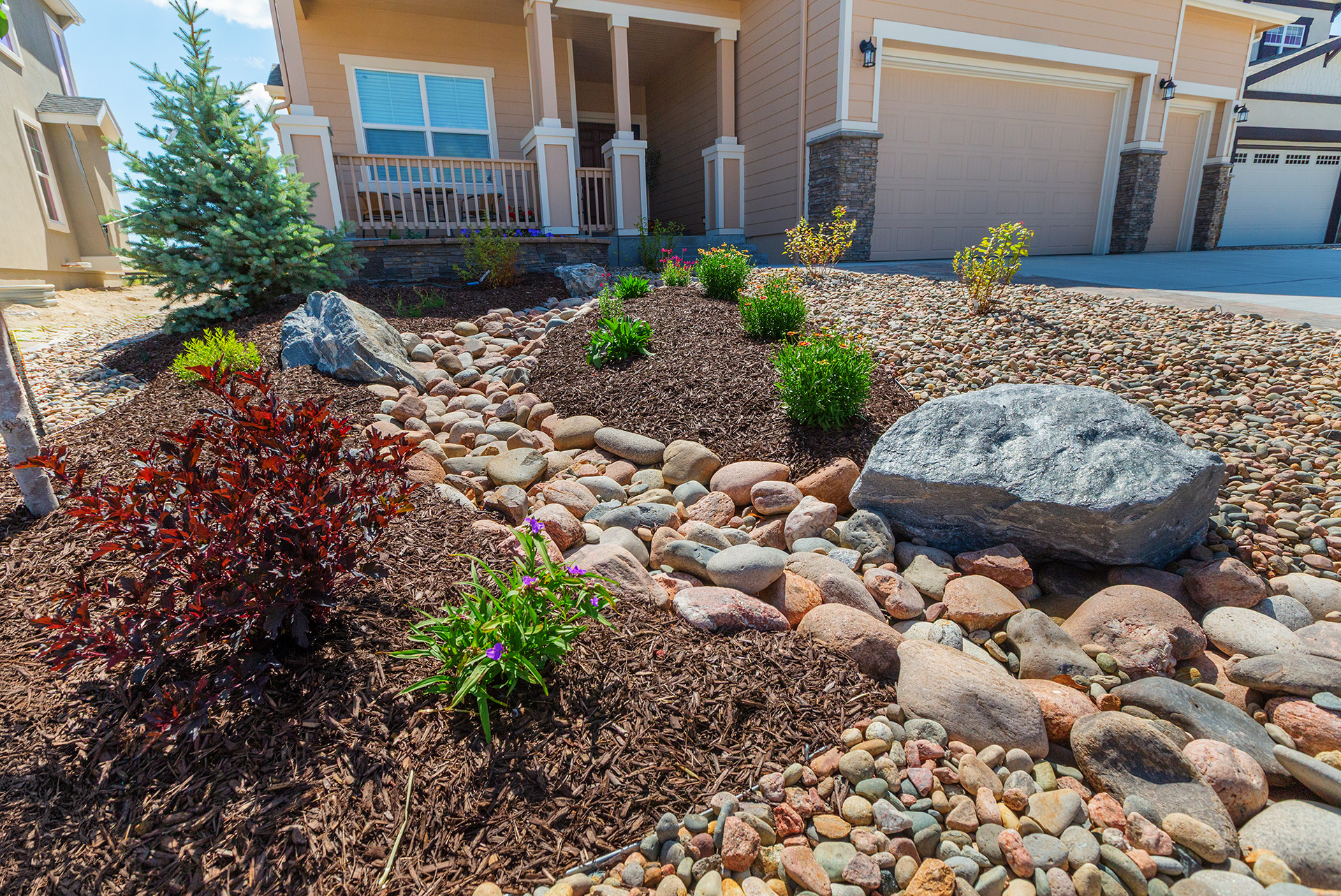 Front yard with xeriscaping, featuring rocks, mulch, small shrubs, and a tree in front of a beige house with a porch and garage.