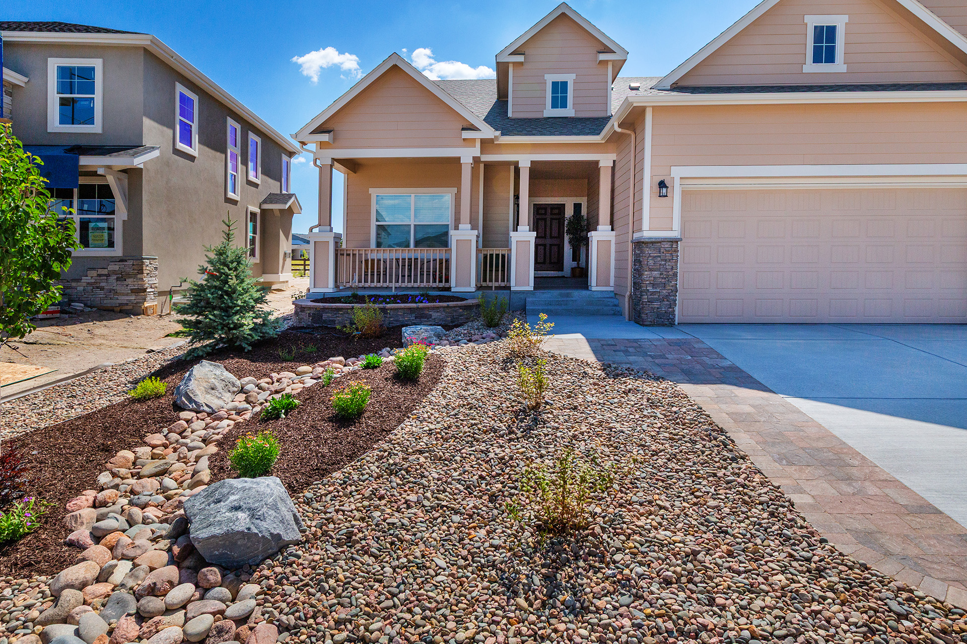 Single-story beige house with a front porch, large garage, and xeriscaped yard featuring rocks, mulch, and small shrubs under a clear blue sky.
