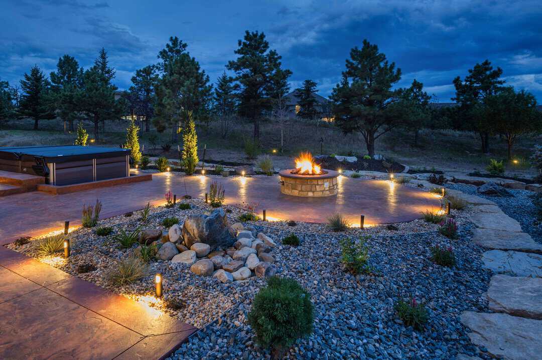 Outdoor patio with a stone fire pit, surrounding landscape lights, rocks, plants, and trees in the background at dusk.