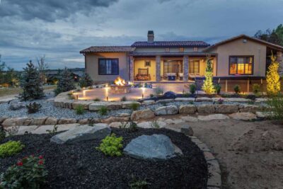 Single-story house with a tile roof, stone patio, and fire pit in a landscaped yard with rocks, shrubs, lights, and trees at dusk.