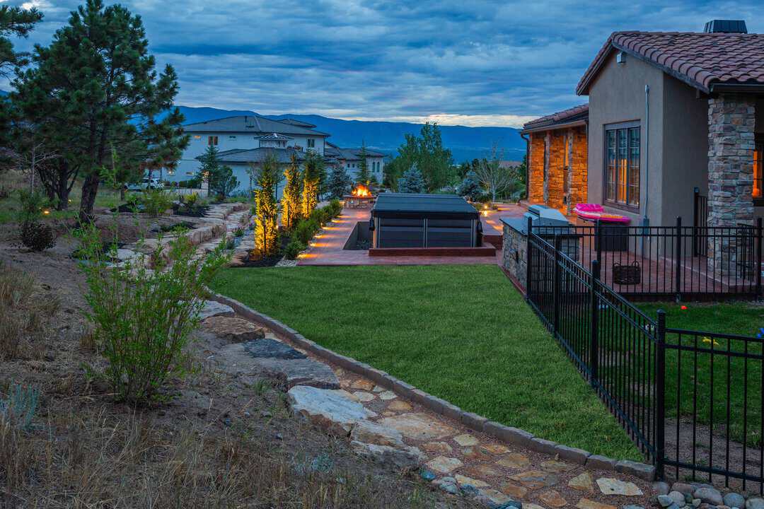 A landscaped backyard with a stone path, green lawn, lit trees, a fenced raised hot tub or pool, and a house with stone accents at sunset.