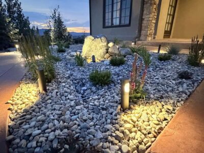 A landscaped front yard with rocks, small plants, outdoor lights, and a large stone near the entrance of a house at sunset.