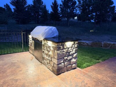 Outdoor stone kitchen island with black countertop and covered grill, illuminated at dusk, on a stamped concrete patio next to a lawn and trees.
