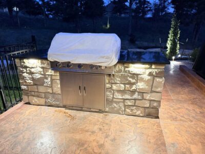 Outdoor stone grill station with a stainless steel grill covered by a white cloth, illuminated by built-in lights on a stamped concrete patio at dusk.