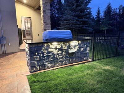 Outdoor grill covered with a tarp, built into a stone counter with lights, next to a fenced yard and patio area in the evening.