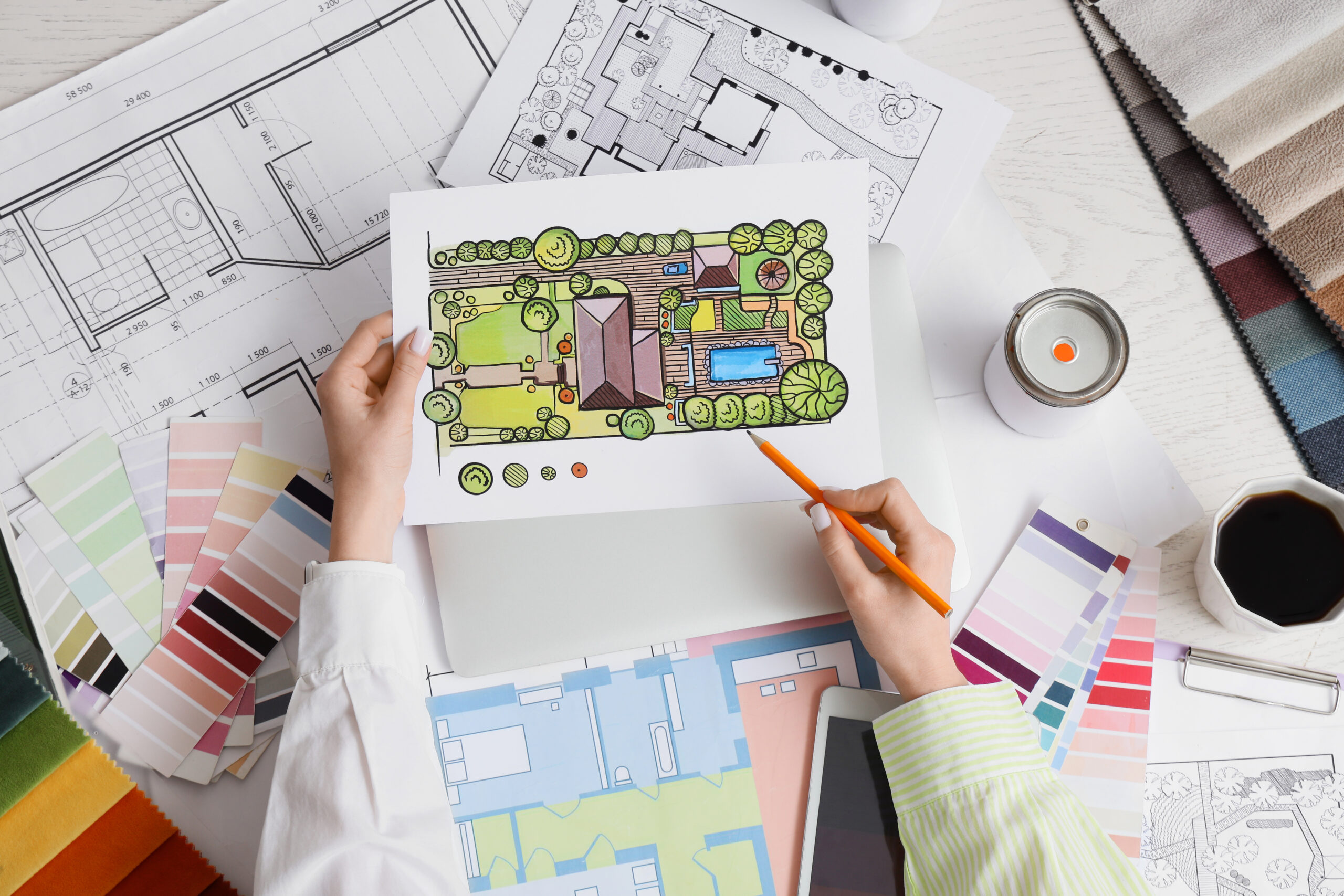 Person holding a colored landscape design plan over architectural blueprints, paint swatches, a candle, and a cup of coffee on a desk.