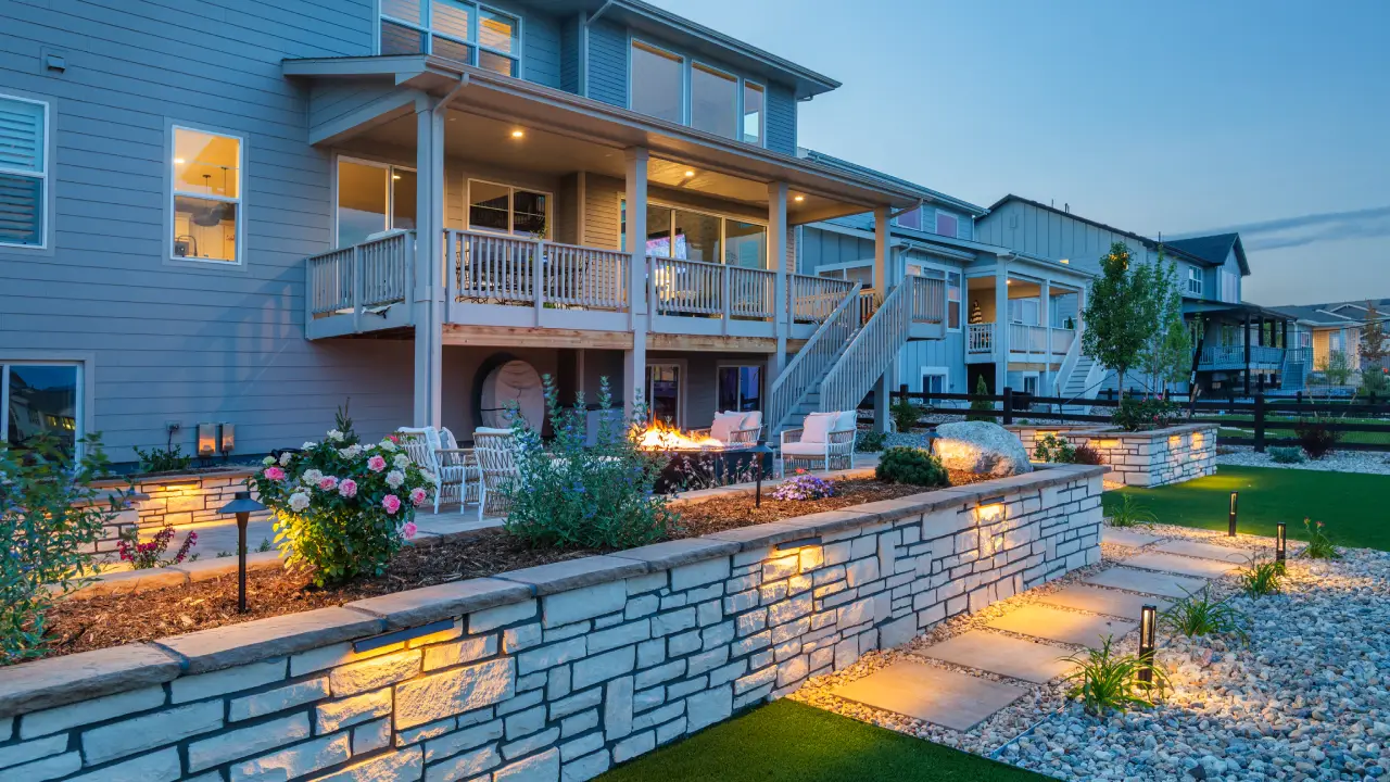 Two-story house with a large back porch, outdoor seating area, fire pit, garden, and illuminated stone retaining wall at dusk.