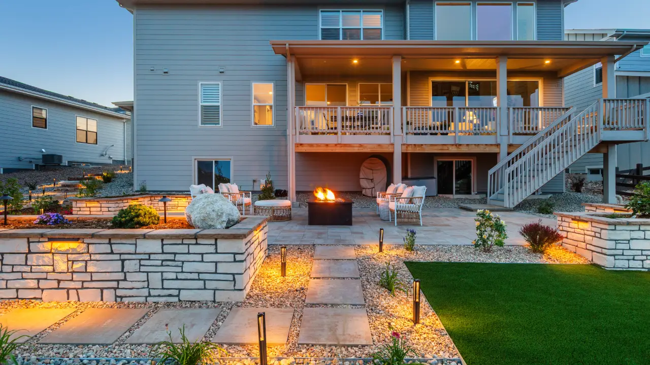 Backyard with stone pathways, a green lawn, illuminated landscaping, seating area, and a fire pit, viewed from behind a modern two-story house with a covered wooden deck.