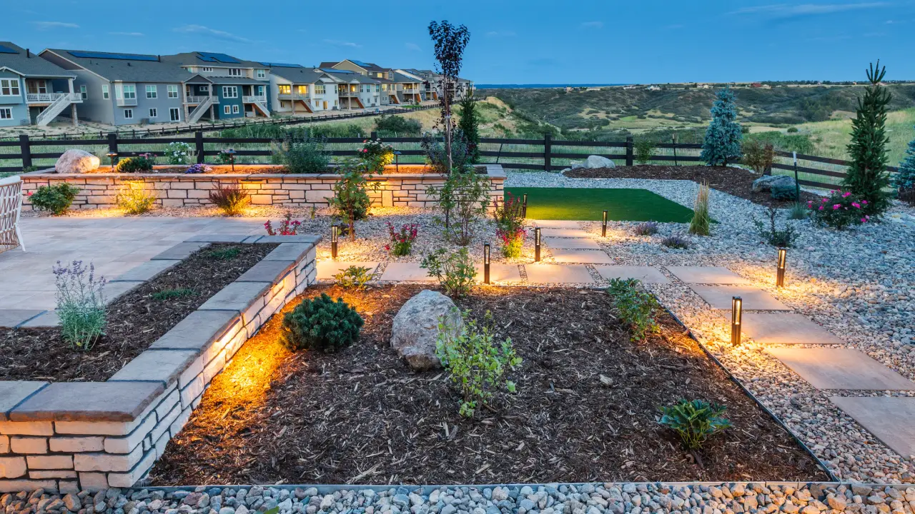 Modern backyard garden at dusk with illuminated pathways, raised flower beds, decorative rocks, artificial turf, and a view of neighboring homes and hills in the background.
