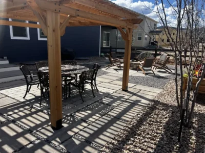 A backyard patio with a wooden pergola, metal dining set, lounge chairs, and gravel landscaping on a sunny day.
